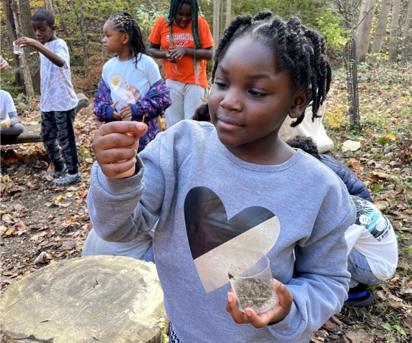 Girl holding a jar of soil in one hand and looking closely at a component of the soil in the other hand.